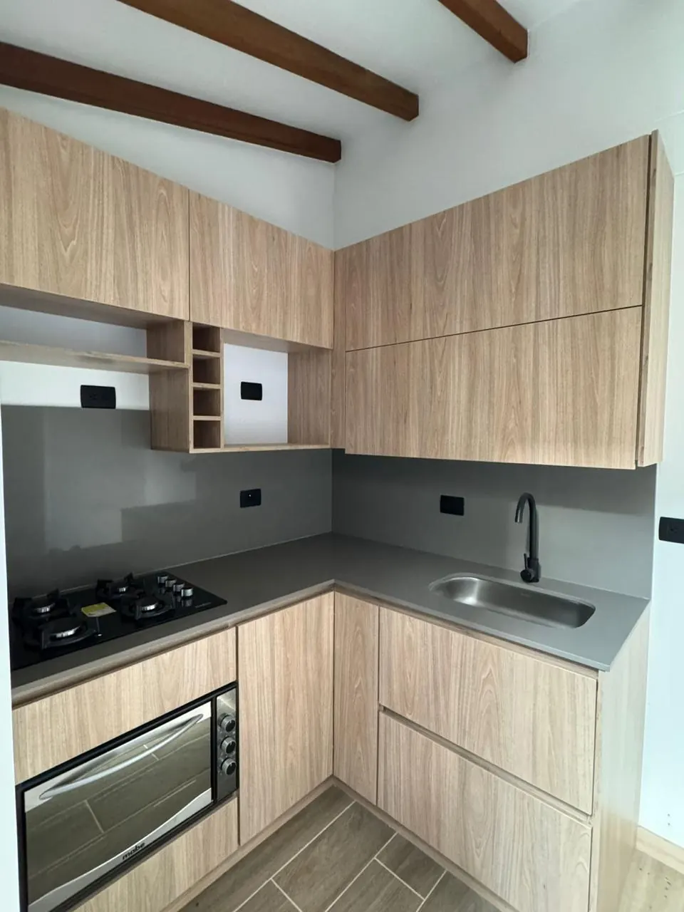 Cocina en L con muebles en madera clara, estufa empotrada, horno y lavaplatos en acero.