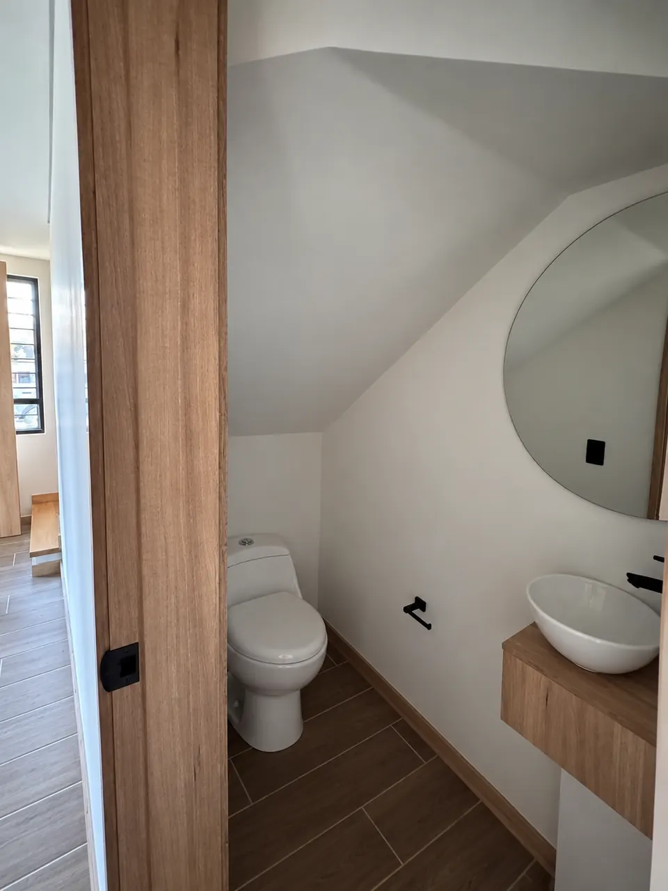 Baño social bajo la escalera con espejo redondo y lavamanos sobre mueble de madera.
