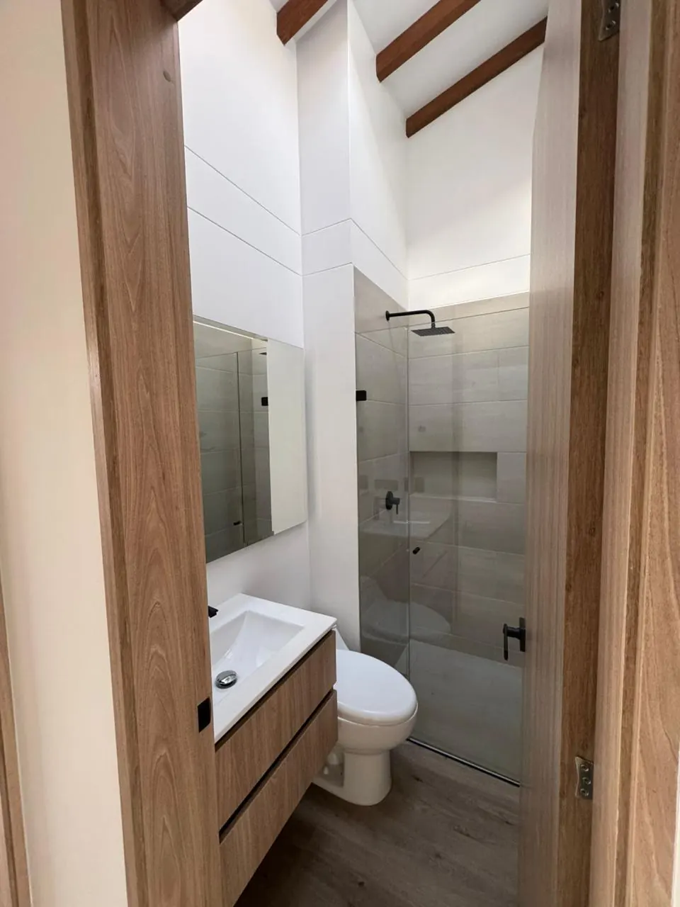 Baño con ducha cerrada en vidrio, grifería negra y techo con vigas de madera.