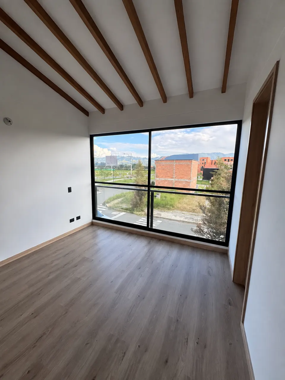 Alcoba con ventanal amplio, techo de vigas de madera y vista a zonas verdes y montañas.